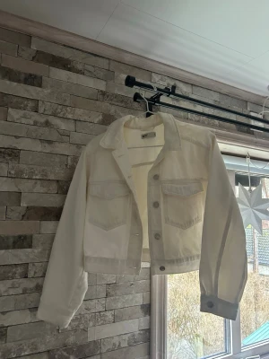 Vit croppad jeansjacka  - Säljer en fräsch vit jeansjacka från Gina Tricot  i croppad modell. Jackan har klassisk krage, två stora bröstfickor med lock och knappar framtill. Materialet är stadigt denim och ärmarna är långa med knapp vid mudden. Perfekt för dig som gillar clean och enkel stil.