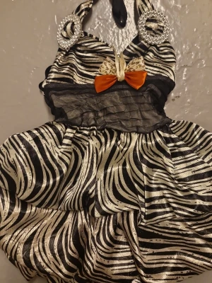 Zebramönstrad kortklänning med rosett - Snygg kortklänning med zebramönster i svart och vitt, dekorerad med en orange rosett och leoparddetalj framtill. Klänningen har pärlprydda ringar vid axelbanden och ett lager av svart. För små flickor och aldrig använt. (Önskar att jag kunde testa den när jag var liten😞)