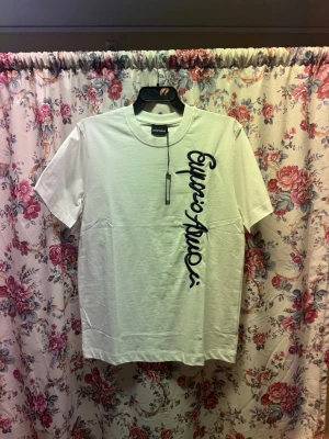 Vit t-shirt från Emporio Armani - Säljer en fräsch vit t-shirt från Emporio Armani med coolt broderat logotyptryck i svart längs framsidan. T-shirten har rund hals och korta ärmar, tillverkad i 100% bomull för skön och mjuk känsla. Perfekt för dig som gillar stilrena designer med en twist.