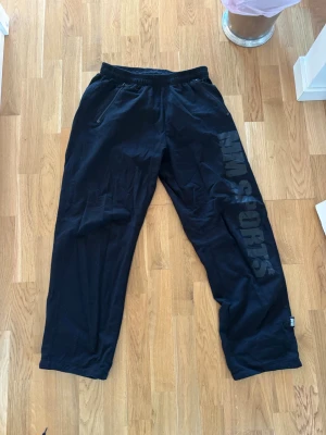 Svarta mjukisbyxor från MM Sports - Svarta mjukisbyxor från MM Sports med tryck längs ena benet. Byxorna har elastisk midja, raka ben och dragkedjeförsedda fickor framtill. Perfekta för chill eller träning, tillverkade i mjukt bomullsmaterial för skön känsla.