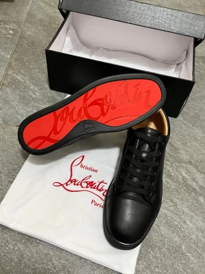 Svarta sneakers från Christian Louboutin - Säljer ett par svarta sneakers från Christian Louboutin med klassisk röd sula och rund tå. Skorna är i skinn med svarta snören och har en stilren, minimalistisk design. Insidan är beige och de levereras med originalpåse och låda.