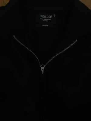 Svart tröja från Indicode - Svart tröja från Indicode med stilren design och silverfärgad dragkedja framtill. Jackan har en klassisk krage, lång ärm och en bröstficka. 