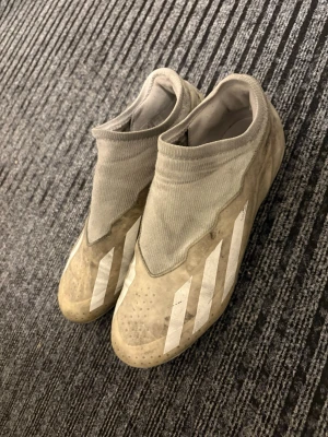 Adidas Predator fotbollsskor - Två par Adidas Predator fotbollsskor säljes. Ena paret är beige med vita ränder och har en strumpliknande ovandel, medan det andra paret är svart och rosa med vita detaljer och mönstrad ovansida. Båda har dobbar för gräsplan och är tillverkade i syntetmaterial.
