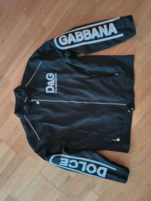 Svart vindjacka från Dolce & Gabbana - Svart vindjacka från D&G med stora vita logotryck fram och bak. Jackan har ståkrage, dragkedja framtill och på fickorna, samt vita detaljer längs axlar och ärmar. Materialet är lätt och syntetiskt, perfekt för en cool streetstil.