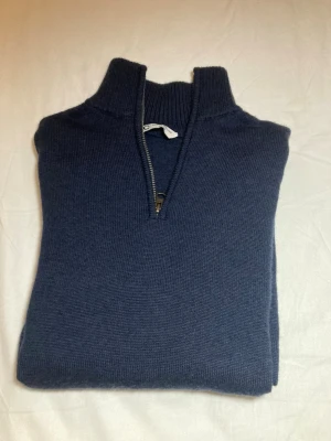 Marinblå merinoulltröja med quarter zip - Säljer en marinblå stickad tröja från Cubus i 100% merinoull. Tröjan har en hög krage med quarter zip-dragkedja och långa ärmar. Ribbstickade muddar vid ärmslut och nederkant ger en snygg passform. Perfekt för en vinter outfit. Tröjan är endast använd några få gånger. Nypris 650-700