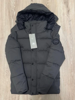 Mörkgrå dunjacka från Canada Goose - Säljer en mörkgrå dunjacka från Canada Goose med huva och svart patch på ärmen. Jackan har quiltad design, dragkedja och knappar framtill. Insidan är fodrad i svart och jackan har en clean, oversized look som är perfekt för kalla dagar.