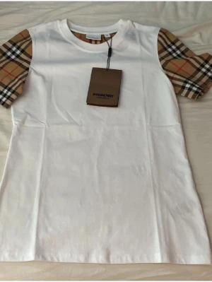 Vit Burberry t-shirt med rutiga ärmar - Säljer en snygg vit t-shirt från Burberry med klassiska rutiga korta ärmar i beige, svart och rött. T-shirten har rund hals och är gjord i mjuk bomull. Perfekt för dig som vill ha en stilren look med en touch av lyx.