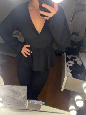 Svart omlottopp med knytband - Säljer en svart omlottopp med långa ärmar och v-ringning. Toppen har knytband i midjan och en lätt volang nedtill som ger en snygg siluett. Köpt från Nelly och aldrig kommit till användning för upplever att den var för stor storlek.
