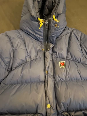 Blå dunjacka från Fjällräven XS - Säljer en blå dunjacka från Fjällräven i storlek XS. Jackan har gul snörning vid kragen, dragkedja och tryckknappar framtill samt Fjällräven-logga på bröstet. Perfekt för kalla dagar med sin fluffiga och quiltade design. Endast använd ett fåtal gånger och bättre bilder på jackan kan skickas ifall efterfrågat! 