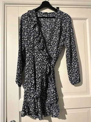 Marinblå blommig omlottklänning från Only - Snygg marinblå omlottklänning med vita blommor, volangkant och knyt i midjan. Klänningen har långärmade ärmar med resår vid mudden och v-ringad hals. Materialet känns lätt och luftigt, perfekt för varmare dagar.