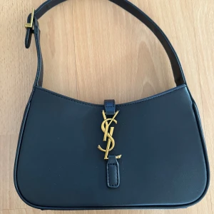 Svart axelväska från YSL - Säljer en stilren svart axelväska från YSL med ikoniskt YSL-emblem i guld på framsidan. Väskan är gjord i slätt skinn och har justerbart axelband.