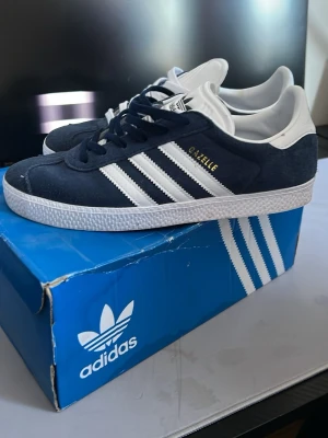 Adidas Gazelle mörkblå sneakers - Säljer ett par Adidas Gazelle sneakers i mörkblå mocka med vita detaljer och klassiska tre ränder på sidan. Skorna har snörning, vit platt sula och rund tå. Adidas-logga på tungan och hälen. Perfekt för dig som gillar retrostil och streetwear.