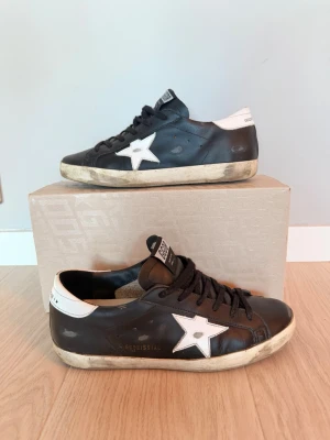 Golden Goose svarta sneakers med vit stjärna - Golden Goose sneakers i svart skinn med vit stjärndetalj på sidan och vit häl med logotyp. Klassisk rund tå, platt sula och svarta snören. Snyggt slitna detaljer och GGDB-märke på plösen. Perfekt för dig som gillar streetstyle och italiensk design.      Skick 8/10