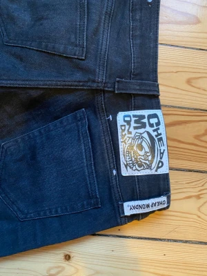 Svarta bootcut Cheap Monday jeans - Säljer ett par svart/mörk gråa jeans från Cheap Monday som är low waist och bootcut och klassisk femficksdesign. Jeansen har en vit patch med grafiskt tryck bak och är gjorda i jeansmaterial med lätt tvättad look. Har några små fläckar, men skulle tro att det går bort i tvätten storlek 26/30 vet att de är lite dåliga bilder så skriv gärna för fler!