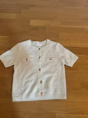 Vit kortärmad kofta från Gina Tricot - Kortärmad vit kofta från Gina Tricot med snygga guldfärgade knappar framtill och på de två dekorativa bröstfickorna. Koftan är stickad och har ribbade kanter vid hals, ärmslut och nederkant. Perfekt för dig som gillar en clean och stilren look. Storlek 146/152. Använd 1-2 gånger, jättebra skick.