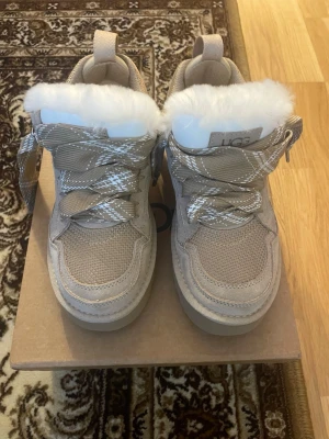 Beige sneakers från UGG med päls - Säljer ett par beige sneakers från UGG med breda mönstrade snören och fluffig vit päls runt öppningen. Skorna har en rund tå, platt sula och är tillverkade i mocka och syntet med meshdetaljer. Perfekta för dig som vill ha både stil och komfort under höst och vinter. Säljer pga de e för små. Är i 40 men känns som 39