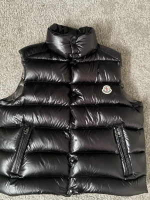 Moncler tibb väst  - En svart Moncler tibb väst i longue saison matrial.  Den är i mycket bra skick då den är obefintligt använd. Den säljs då den är för liten och storleken är 2(M). Västen är dessutom varm och utmärkt både till vintern och framåt våren. På moncler hemsida beskrivs västen följandeA Moncler classic, the Tibb gilet for men revisits the archives and draws inspiration from iconic silhouettes of the 80’s. Perfect for the mid-season. På grund av flytt välkomnas samtliga prisförslag. Vi hörs!