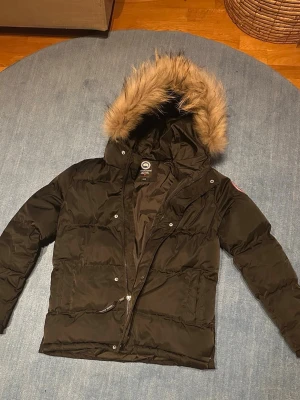Svart Canada Goose dunjacka med päls - Säljer en helt ny, oanvänd svart dunjacka från Canada Goose med fluffig päls (äkta) på huvan och klassisk logga på ärmen. Jackan har dragkedja, knappar och två fickor framtill. Perfekt för kalla dagar och har en riktigt snygg look. Står att det är 15-16 år men kan även passa yngre barn beroende på storlek. Min 12-åring provade och den var bara lite för stor för honom och han är normalbyggd.