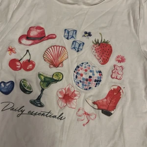 Vit t-shirt med färgglada tryck från FASher - Vit t-shirt från FASher i storlek S med coola och färgglada tryck på framsidan, bland annat cowboyhatt, körsbär, lime, discokula, blomma och cowboy boots. T-shirten har rund hals och korta ärmar. Materialet är mjuk bomull och passformen är normal.