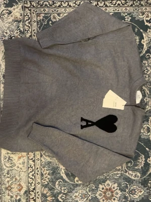 Grå stickad tröja från Ami - Säljer en grå stickad tröja från Ami Alexandre Mattiussi med svart hjärta och bokstaven A broderat på bröstet. Tröjan har rund hals, ribbade muddar och lång ärm. Perfekt för dig som gillar stilrena detaljer och vill ha något som sticker ut.