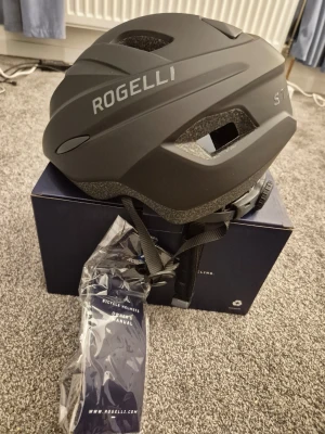 Svart cykelhjälm Rogelli Start - Den är helt ny. Snygg svart cykelhjälm från Rogelli, modell Start. Hjälmen har en matt finish, justerbart spänne bak och flera ventilationshål för bra luftflöde. Passar perfekt för cykling och har en modern, sportig look.