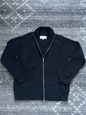 Svart stickad kofta från Maison Margiela - Svart stickad kofta från Maison Margiela med hel dragkedja framtill och ribbade muddar. Koftan har en stilren design med hög krage och klassiska Margiela-stygnen i nacken. Perfekt för dig som gillar minimalistisk och cool stil.