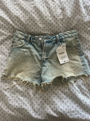 Lågmidjade Jeansshorts från ZARA - Säljer ett par ljusblå denimshorts från ZARA i storlek 32. De har fransig kant, slitna detaljer och klassisk femficksdesign. Shortsen ärlowwaist och har knapp och dragkedja framtill. Perfekta för en chill sommardag. De är helt oanvända, nypris 329kr