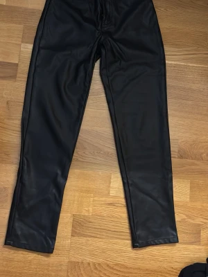 Svarta skinnbyxor med rak passform - Säljer ett par svarta skinnbyxor med rak passform. Byxorna har en clean look utan synliga detaljer och är tillverkade i ett syntetiskt skinnmaterial som ger en cool vibe. Perfekta för dig som vill ha en edgy stil.