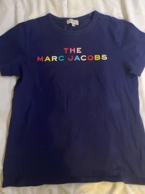 Blå t-shirt från Marc Jacobs - Snygg marinblå t-shirt från Marc Jacobs med färgglad text på bröstet. Klassisk rund halsringning och korta ärmar. Perfekt för dig som vill ha en enkel men ändå statement t-shirt i garderoben.