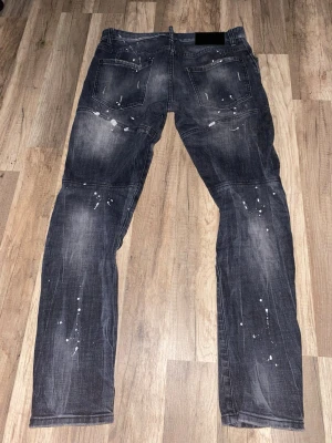Svarta Dsquared2 jeans med slitningar - Säljer ett par svarta jeans från Dsquared2 med coola slitningar och vita färgstänk. Jeansen har en slim passform och klassisk femficksdesign. Materialet är jeans och de har en mörk tvätt med slitna detaljer framtill för en edgy look.