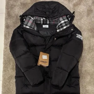 Svart pufferjacka från Burberry - Svart pufferjacka från Burberry med stor huva och rutig innerfoder i grått, vitt och rött. Jackan har en tydlig Burberry London England-patch på ärmen och dragkedja framtill. Perfekt för kalla dagar och har en riktigt schysst streetstil.