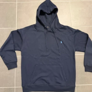 Mörkblå hoodie från Polo Ralph Lauren - Mörkblå hoodie från Polo Ralph Lauren med klassisk huva och snörning. Tröjan har en broderad blå logga på bröstet och en stor känguruficka framtill. Tillverkad i mjuk bomull och har ribbade muddar vid ärmslut och nederkant.