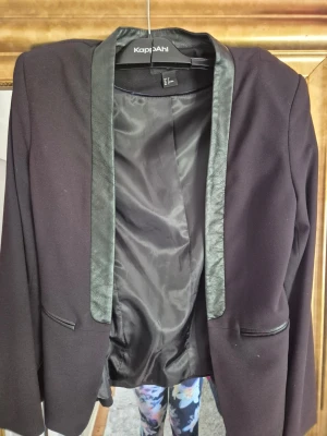 Svart kavaj med skinnkrage från HM - Svart kavaj från HM med snygga detaljer i skinn på kragen och fickorna. Klassisk rak passform och långärmad design. Perfekt för dig som gillar stilrena plagg med en liten edge. Materialet är mjukt och känns bekvämt mot kroppen.