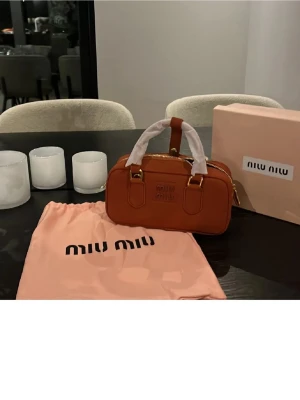 Brun handväska från Miu Miu - Säljer en snygg brun handväska från Miu Miu i skinn med guldfärgade detaljer och dragkedja. Väskan har ett stilrent, rektangulärt format och två handtag. Perfekt storlek för det viktigaste och kommer med original dustbag och box.