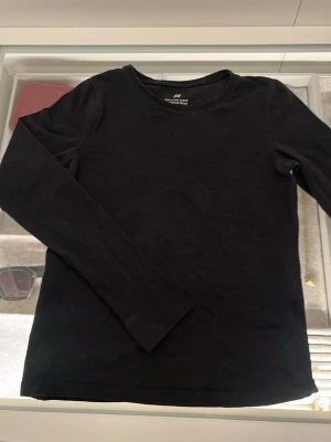 Svart långärmad topp från H&M - En enkel svart långärmad topp från H&M i storlek 134/140. Modellen har rund hals och är tillverkad i mjuk bomull som känns skön mot huden. Perfekt att styla med jeans eller under en hoodie. Snygg och stilren basic som funkar året runt.