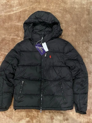 Svart pufferjacka från Polo Ralph Lauren - Svart pufferjacka från Polo Ralph Lauren med huva och flera dragkedjeförsedda fickor. Jackan har det klassiska röda Polo-logot broderat på bröstet och är tillverkad i polyester. Perfekt för kalla dagar och har en clean, sportig vibe.