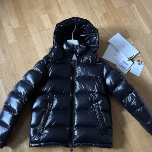 Svart pufferjacka från Moncler - Säljer en svart, glansig pufferjacka från Moncler med huva och två sidofickor med dragkedja. Jackan har en quiltad design och synlig logga på ärmen. Perfekt för kalla dagar och har en riktigt snygg streetstil.