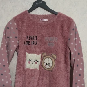 Rosa fleece-tröja med prickar och motiv - Supermjuk rosa fleece-tröja med långärmade ärmar i prickigt mönster. Framsidan har gulliga applikationer av en kudde och en väckarklocka samt texten 'Pleaseee come back!'. Perfekt för mysiga dagar hemma.