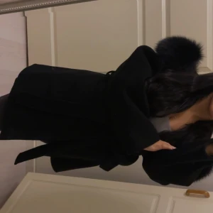 Svart kappa med äkta päls  och bälte - Säljer en svart kappa med bred krage i äkta päls  och knytskärp i midjan. Jackan har lång ärm och en oversized, loose passform. Perfekt för dig som vill ha en stilren och trendig look med extra lyxig känsla.