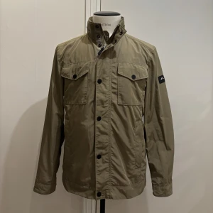 J Lindeberg Grön Field Jacket - Snygg grön vindjacka från J.Lindeberg med hög krage, ihopfällbar huva och svarta knappar. Jackan har två bröstfickor med lock och knäppning samt justerbara detaljer i midjan. Perfekt för dig som gillar stilren och funktionell design. Jackan har några små mindre fläckar framtill men inget som knappt syns eller något man lägger märke till.