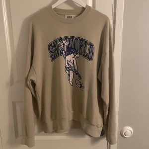 Beige SNS World sweatshirt med tryck - Säljer en beige sweatshirt från SNS med stort blått tryck framtill som föreställer en ängel och fjärilar samt texten 'SNS WORLD'. Tröjan har rund hals, långa ärmar och ribbade muddar. Perfekt för dig som gillar streetwear vibes.