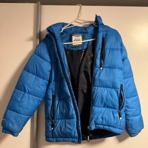 Blå pufferjacka från McKinley - Blå pufferjacka från McKinley med svarta detaljer och dragkedjor. Jackan har huva, tre fickor med dragkedja och quiltad design. Perfekt för kalla dagar och enkel att matcha med streetwear eller casual outfits.