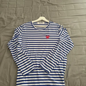 Randig långärmad tröja Comme des Garçons - Säljer en blåvit randig långärmad tröja från Comme des Garçons Play. Tröjan har ett ikoniskt rött hjärta med ögon broderat på bröstet och är gjord i mjuk bomull. Jättebra skick och väldigt nice tröja!