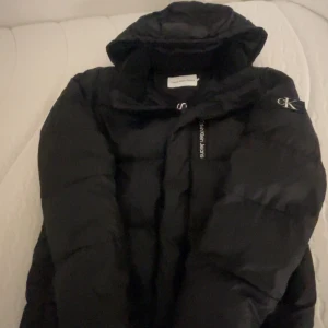 Svart pufferjacka från Calvin Klein Jeans - Säljer en svart pufferjacka från Calvin Klein Jeans med huva och stor logga längs dragkedjan. Jackan har en clean look med CK-märke på ärmen och är perfekt för kalla dagar. Oversized passform och riktigt skön att bära.