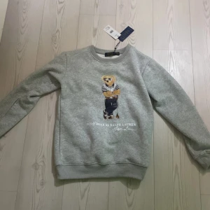 Ralph lourent tröja - Säljer en stilren grå sweatshirt från Polo Ralph Lauren med en ikonisk björn på framsidan. Tröjan har långa ärmar och ribbade muddar vid ärmslut och nederkant. Perfekt för en avslappnad look.