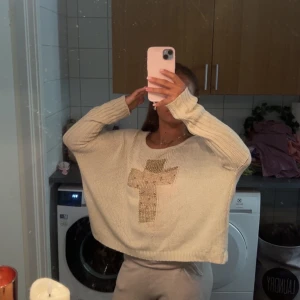Beige stickad tröja med paljettkors - Säljer en beige stickad tröja med ett stort kors i guldiga paljetter på bröstet. Tröjan har loose passform, breda ärmar och rund halsringning. Perfekt för dig som gillar statement-detaljer och chill vibe.