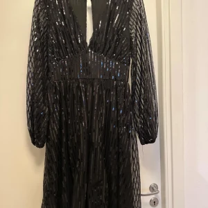 Svart glittrig långklänning från NLY Eve - Svart långklänning från NLY Eve med glittriga paljetter och djup v-ringning. Klänningen har långa transparenta ärmar och ett snyggt band i midjan. Materialet är polyester med paljettmönster som ger extra bling. Perfekt för dig som vill sticka ut och glänsa.
