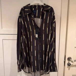 Mönstrad blå skjorta från Gant - Snygg skjorta från Gant i mörkblått med vertikala ränder och guldfärgade nyckelmönster. Skjortan har klassisk krage, knäppning framtill och långa ärmar. Perfekt för dig som vill sticka ut med ett coolt mönster och stilren look.