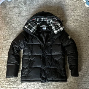 Svart pufferjacka från Burberry - Svart pufferjacka från Burberry med huva och rutigt foder i svart, vitt och lite rött. Jackan har dragkedja och knappar framtill, två fickor med dragkedja och en snygg Burberry-logga på ärmen. Materialet är polyester och jackan har en loose passform.