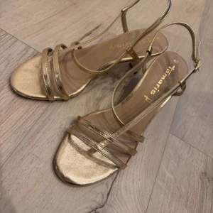 Guldiga sandaler från Tamaris - Snygga sandaler i metallic guld från Tamaris med smala remmar och öppen tå. Skorna har låg klack och justerbar rem runt hälen. Perfekta för dig som vill sticka ut med en glansig look. Passar till både jeans och klänning.